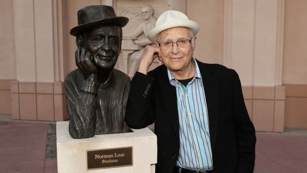 A man mimics a statue of himself leaning his head against his right hand. Statue and man both wear the same hat. The statue is on a pedestal with a plaque that says, "Norman Lear Producer."